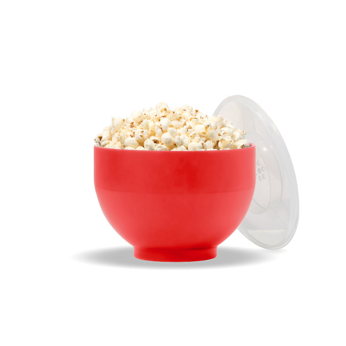 A W&P Reusable Popcorn Popper in red, brimming with popcorn, rests on a white surface with its clear lid leaning casually against it.