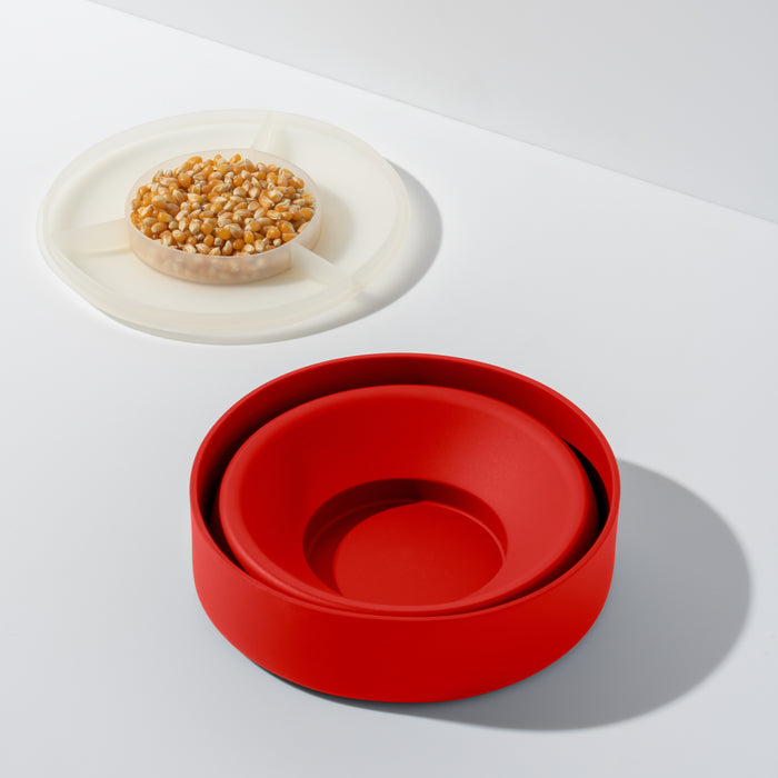 A red circular Reusable Popcorn Popper by W&P with layered sections rests open on a white surface, ideal for holding popcorn. Nearby, a translucent container filled with yellow kernels shines. Shadows cast to the right. Both containers are dishwasher-safe for added convenience.