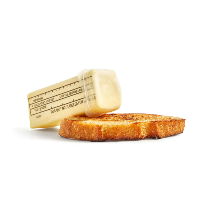 A W&P Reusable Produce Saver Stretch Lid sits beside a clear measuring jar filled with butter, lying sideways against a slice of toasted bread. The jar shows tablespoon and teaspoon markings, all on a plain white background.
