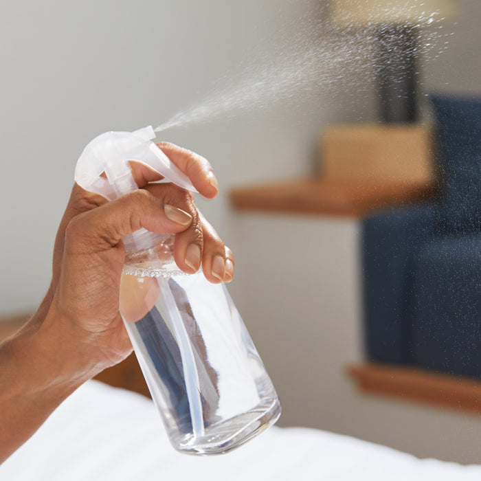 A hand sprays a clear liquid from a Grove Co. Reusable Room Spray Bottle against a blurred room interior backdrop with a bed and wooden furniture. Droplets linger, highlighting this sustainable solution for refreshing your space.