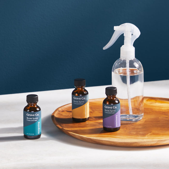 Three Grove Co. Reusable Room Spray bottles rest on a tray next to another transparent version, against a deep blue wall. This setup provides an elegant and sustainable air freshener alternative.