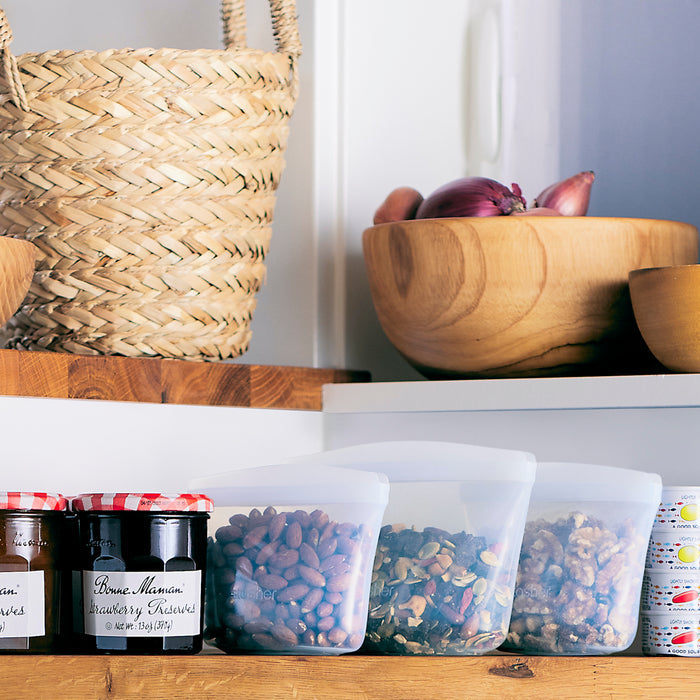 A kitchen shelf displays jam jars, labeled bottles, and three nut-filled translucent containers. For easy lunch prep, a Stasher Reusable Silicone Bowl holds onions and garlic above, with a woven basket nestled in the corner.