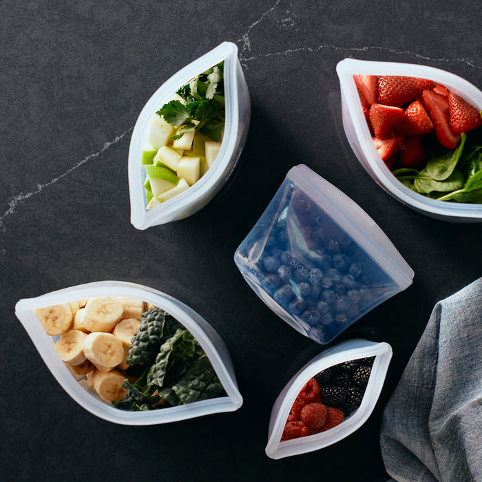 Five reusable silicone bowls from the Stasher Variety Pack hold strawberries, spinach, blueberries, mixed berries, banana slices with kale, and apple cubes with parsley on a dark surface. A gray cloth is partially visible. Ideal for lunch prep!.