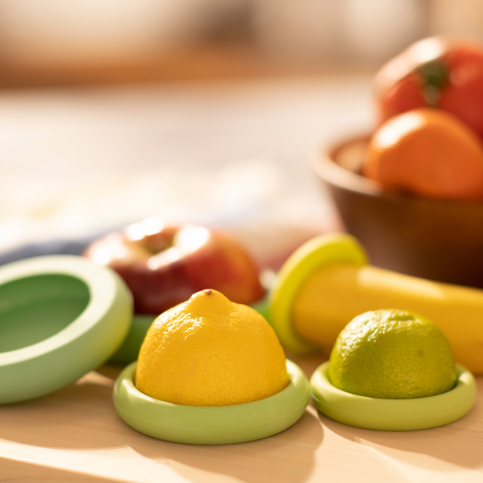 A lemon and a lime sit in two green Reusable Silicone Food Savers by Food Huggers on a wooden table, highlighting fresh produce preservation. In the blurred background, a bowl contains apples and oranges. The brightly lit scene suggests a sunny environment filled with natural light.