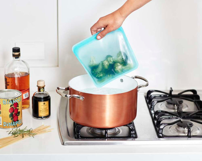 A hand is adding broccoli from a BPA-free, reusable Stasher Silicone Sandwich Bag into a copper pot of boiling water on the stove. Nearby are spaghetti, a can of tomatoes, balsamic vinegar, and olive oil for a simple yet flavorful meal.