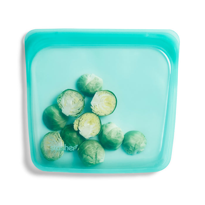 A teal, BPA-free Stasher Reusable Silicone Sandwich Bag holds Brussels sprouts, some halved. Its square design with rounded edges and a clear surface highlights fresh contents, perfect for eco-friendly food storage.