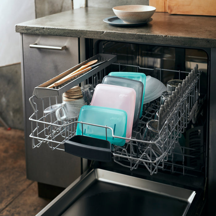Open dishwasher with a loaded rack containing Stasher brand Reusable Silicone Snack Bags, jars, and utensils. The kitchen features concrete countertops and dark cabinetry, while a wooden cutting board and stacked bowls are on the counter.