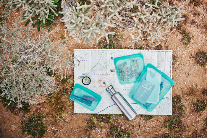 A Stasher Reusable Silicone Snack Bag and a non-toxic, metal water bottle are placed on a topographic map, surrounded by desert foliage, suggesting readiness for an eco-friendly outdoor adventure.