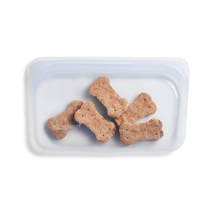 Five bone-shaped, non-toxic dog treats are randomly packed in a temperature-resistant, transparent rectangular Reusable Silicone Snack Bag. Made from premium food-grade silicone and branded with Stasher, these uniformly brown textured treats are neatly sealed inside.