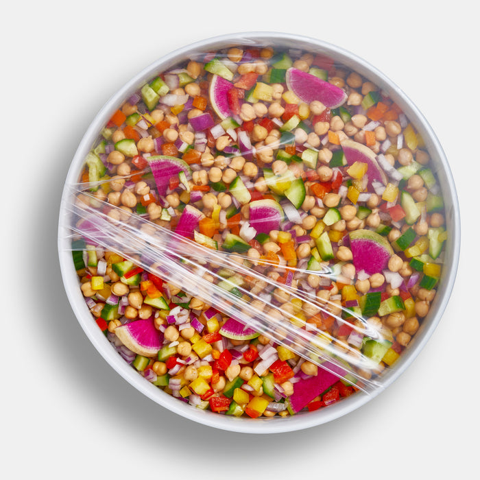 A colorful salad with chickpeas, diced cucumbers, red onions, red bell peppers, and sliced radishes is neatly covered by a W&P Reusable Silicone Stretch Wrap on a white background, highlighting an eco-friendly plastic alternative.