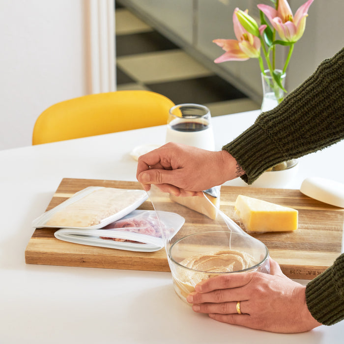 A person wearing a green sweater covers a glass bowl with W&Ps Reusable Silicone Stretch Wrap on a wooden cutting board. Nearby are cheese blocks, deli meat in a container, and a vase of pink lilies, with a yellow chair visible in the background.