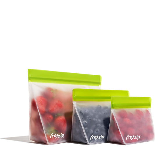 Three (re)zip Reusable Stand-Up Bags made of food-grade PEVA, each with a green zip, hold strawberries, blueberries, and raspberries. Semi-transparent and leakproof, they are displayed in a row.