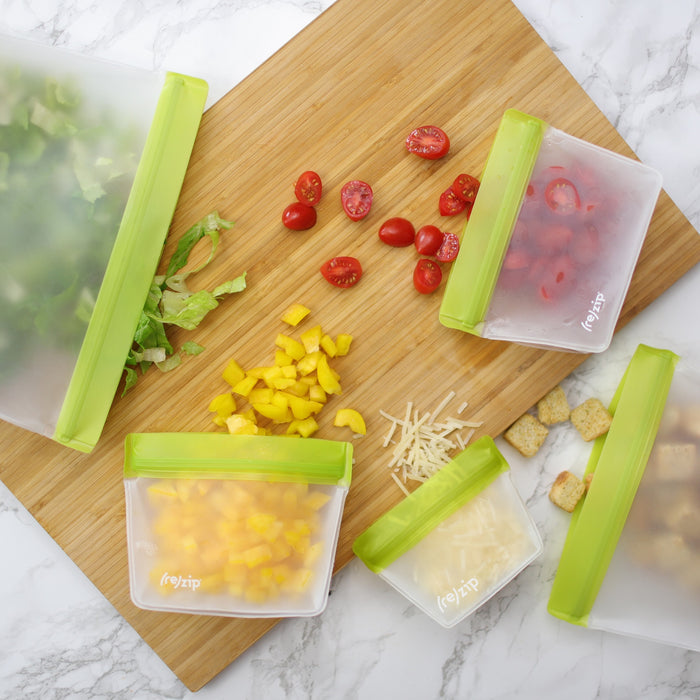 Zip Reusable Stand-Up Bags with green seals, made from food-grade PEVA, hold chopped lettuce, cherry tomatoes, bell peppers, cheese, and croutons on a wooden cutting board with extra veggies nearby.