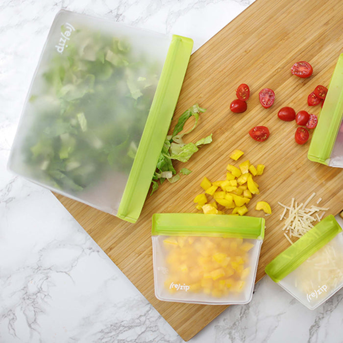 Three (re)zip Reusable Stand-Up Bags with green seals, made from food-grade PEVA, store chopped lettuce, yellow bell pepper, and shredded cheese on a wooden cutting board with sliced cherry tomatoes and extra yellow pepper.