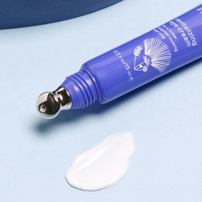 Close-up of Cocokinds Revitalizing Eye Cream in a blue tube with a metal applicator, ideal for dark circles and wrinkles, on a light blue surface. A small dab of white cream is beside it.