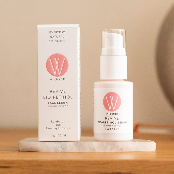 A bottle of Wildcrafts Revive Bio-Retinol Face Serum, with pink and white packaging, featuring plant-based retinol alternatives like rambutan and evening primrose, rests beside its box on a stone base. Set on wood with soft lighting, it promises revitalized skin.