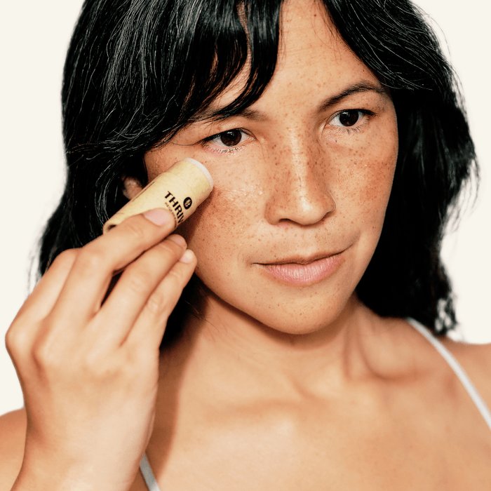 With long, dark hair and freckles, the person applies Thrive Natural Cares Revive Eye Stick to their cheek. Its natural and minimalist design aligns with their focused demeanor, offering bakuchiol benefits for radiant skin.