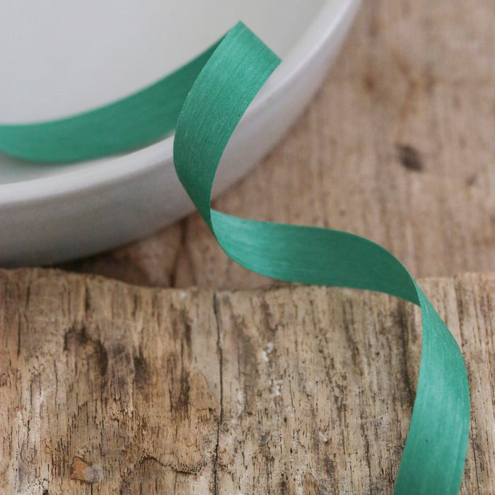 A teal Ribbon from Waterleaf Paper Company, made of Wisconsin cotton, beautifully curls on a rustic wooden surface next to a white bowl. The textured wood contrasts with the smoothness of the sustainable ribbon and bowl, forming a captivating composition.
