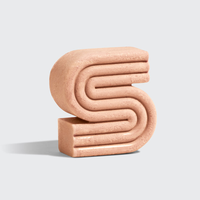 The Rice Water Protein Shampoo Bar by Kitsch features a sculptural, abstract S design in a smooth, stone-like material against a light gray background. Inspired by rice waters flow, its rounded, layered elements create a three-dimensional effect.