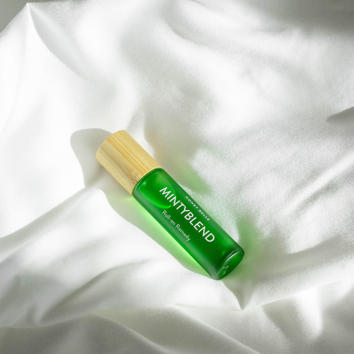 Resting on a softly textured white fabric, Honey Belles Roll-on Remedy features a green bottle with a bamboo cap labeled MINT BLEND. Illuminated by gentle natural light, it casts subtle shadows and is perfect for soothing aromatherapy with its essential oils.