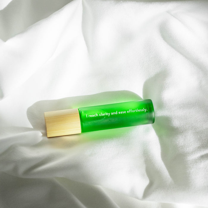 A green Honey Belle Roll-on Remedy bottle with a wooden cap rests on white crumpled fabric. Sunlight creates soft shadows around it, illuminating the text: I reach clarity and ease effortlessly. Its infused with essential oils.