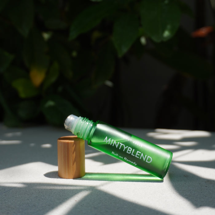 A green Roll-on Remedy by Honey Belle, with a wooden cap, lies open on a light surface. Sunlight casts leaf shadows, creating a dappled effect against the lush, blurred green foliage background, evoking soothing aromatherapy.