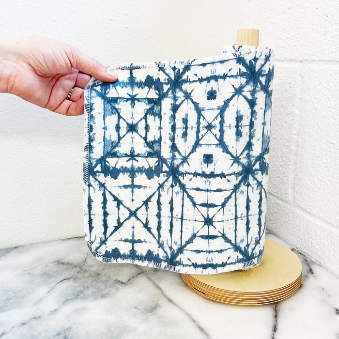 A hand holds a blue and white patterned Rolled Unpaper® Towel from Marleys Monsters, partially unrolled from a wooden holder on a marble surface against a white wall. These eco-friendly towels are perfect for sustainable living.