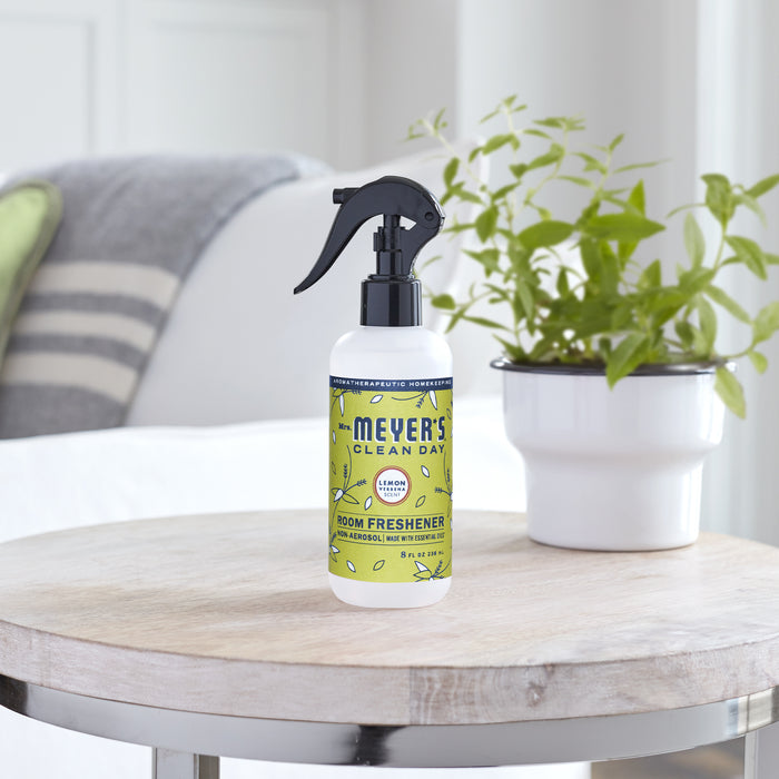 A bottle of Mrs. Meyers Room Freshener with a black nozzle rests on a round wooden table. The green label, adorned with lemon illustrations, suggests a hint of essential oils inside. A white pot with green leaves and a gray-and-white striped pillow in the background add to the fresh ambiance.