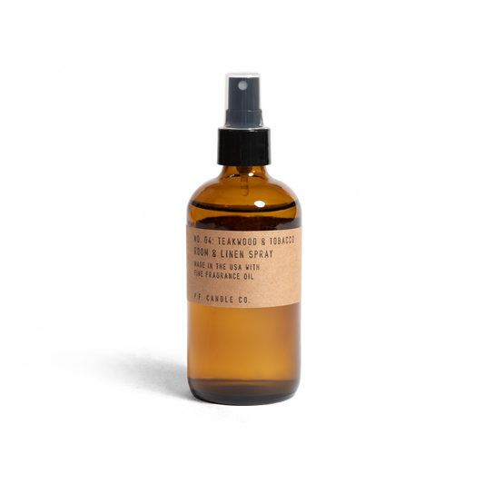 A brown glass spray bottle labeled No. 04: Teakwood & Tobacco Room & Linen Spray by P.F. Candle Co., crafted in the USA with fine fragrance oil for a rich woody scent, features a sleek design with a black spray nozzle.