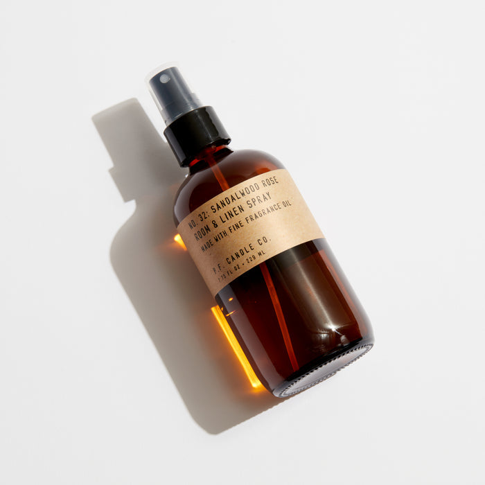 The P.F. Candle Co. Room & Linen Spray in sandalwood rose features a woody scent and a pump nozzle. It comes in a brown bottle with a simple label, set against a plain light backdrop that elegantly casts shadows to the left.