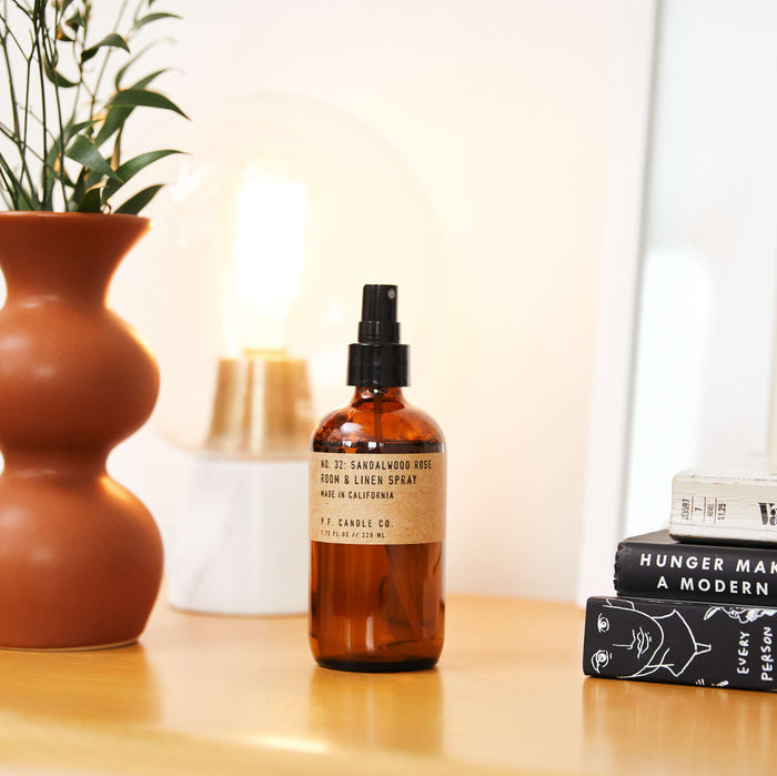 A P.F. Candle Co. Room & Linen Spray in a brown glass bottle labeled Sandalwood Rose sits on a wooden surface near books and a ceramic vase with leaves. A soft, glowing lamp enhances the woody scent, creating a cozy atmosphere.