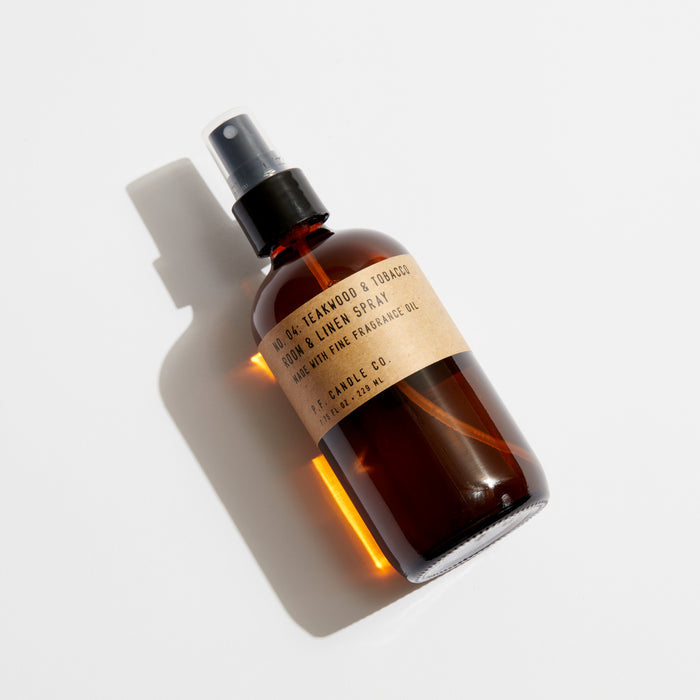 A P.F. Candle Co. Room & Linen Spray in a dark amber bottle labeled Cedar, Teakwood & Tobacco sits on a white surface, exuding its woody scent. Black text adorns the label, while the bottle casts a subtle shadow to the left.