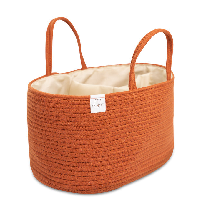The Natemia Rope Diaper Caddy, a medium oval-shaped nursery basket made from orange GOTS Certified Organic Cotton, features two handles, a white interior, and a small tag with a smiley face. It’s perfect for use as a baby nappy caddy.