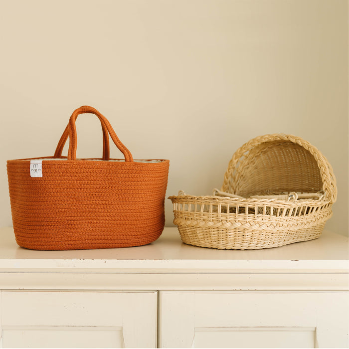Displayed on a white cabinet with a cream background are two baskets: an orange one with handles for nursery storage and the Natemia Rope Diaper Caddy, featuring a light-colored woven style in GOTS Certified Organic Cotton with an open top.