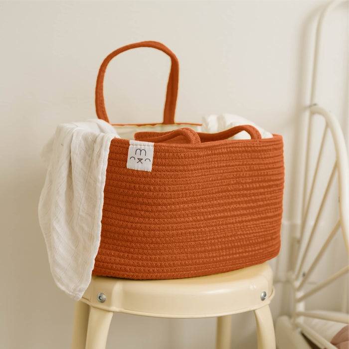 A Natemia Rope Diaper Caddy, ideal for nursery storage, rests on a cream chair. A white blanket cascades over its side, enhancing its charm. The backdrop is a gentle, light-hued wall that completes this cozy scene.