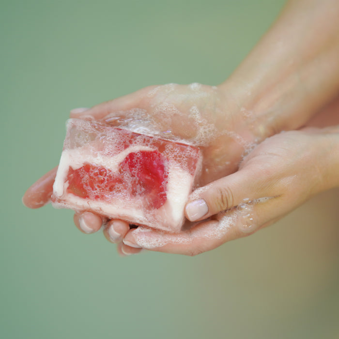 Hands cradle the Blooming Rose Soap Bar by Soapymania, translucent with red and white swirls covered in bubbles, offering a moisturizing experience. Crafted from natural handmade ingredients, it rests against a soft, blurred green backdrop.