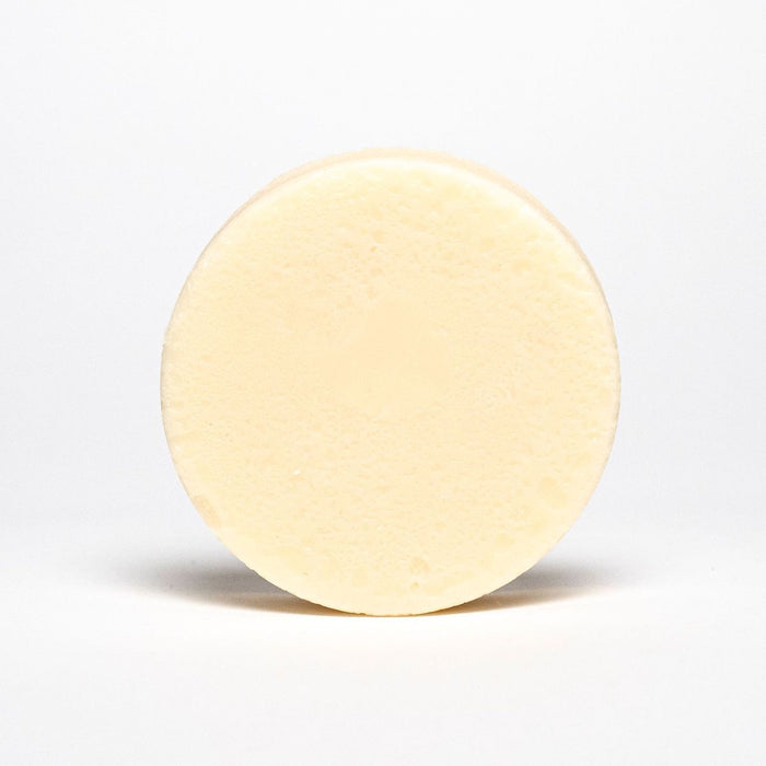 The Rosemary Mint Conditioner Bar by Spinster Sisters Co. is a circular, solid cream-colored sulfate-free bar with a smooth, slightly textured surface. It stands upright against a white background, embodying plastic-free and Fair Trade principles.
