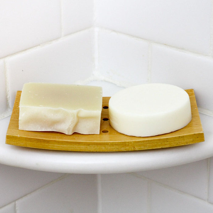 Two Spinster Sisters Co. Rosemary Mint Conditioner Bars sit on a wooden dish in the bathroom corner, one rectangular and textured, the other smooth and circular. The white tiled walls perfectly highlight these plastic-free, sulfate-free bars.