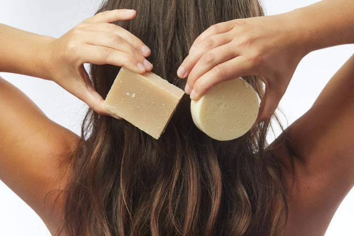 A person with long brown hair holds a Spinster Sisters Co. Rosemary Mint Conditioner Bar in one hand and a round bar of soap in the other, set against their head. The background is plain white.