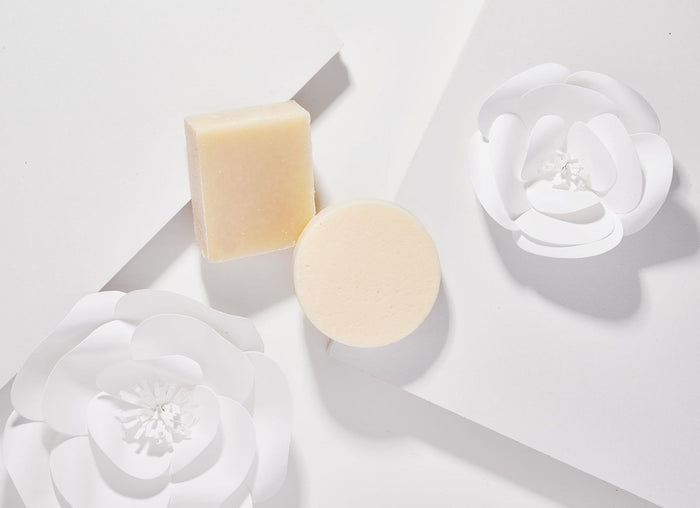 A square and a round bar of soap lie on a white background, surrounded by white paper flower cutouts. The minimalistic setting highlights the soaps natural tones and the Rosemary Mint Shampoo Bar from Spinster Sisters Co., enriched with essential oils and free of sulfates.