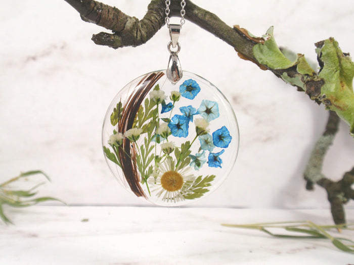 Round crystal resin pendant showing blue flowers and greenery with a lock of hair inside.