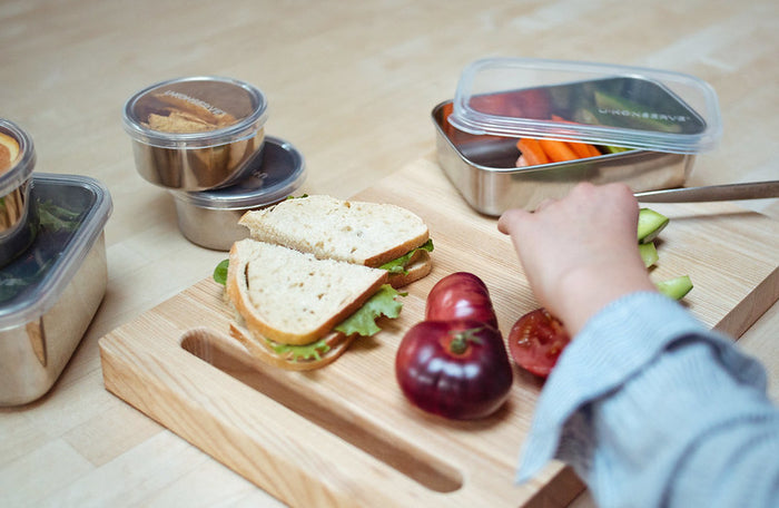 A person arranges a packed lunch on a wooden board with a sandwich, apple, and cut vegetables stored in U-Konserves Round Medium Container. This setup showcases a dedication to healthy eating and plastic-free living.