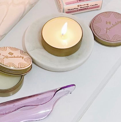 A lit ThreePlay Massage Candle by Lovability, in a round tin, sits on a white marble coaster. Its surrounded by closed tins with decorative lids and accompanied by a box labeled ThreePlay Massage Candle and a translucent purple object, all placed on a light surface.