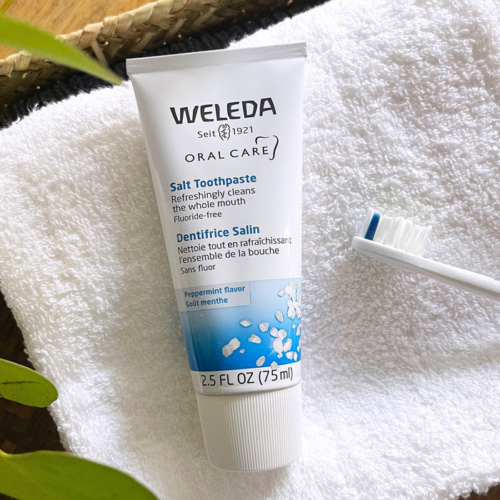 Weleda Salt Toothpaste, featuring blue and black accents on its packaging, sits on a white towel. Surrounding it are a green leaf and a white toothbrush, evoking the fresh feeling of peppermint.