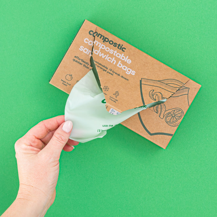 A hand opens a Compostic Sandwich Bags box made from recycled materials on a green background, revealing a light green, home-compostable bag with sandwich illustrations crafted from plant-based materials.