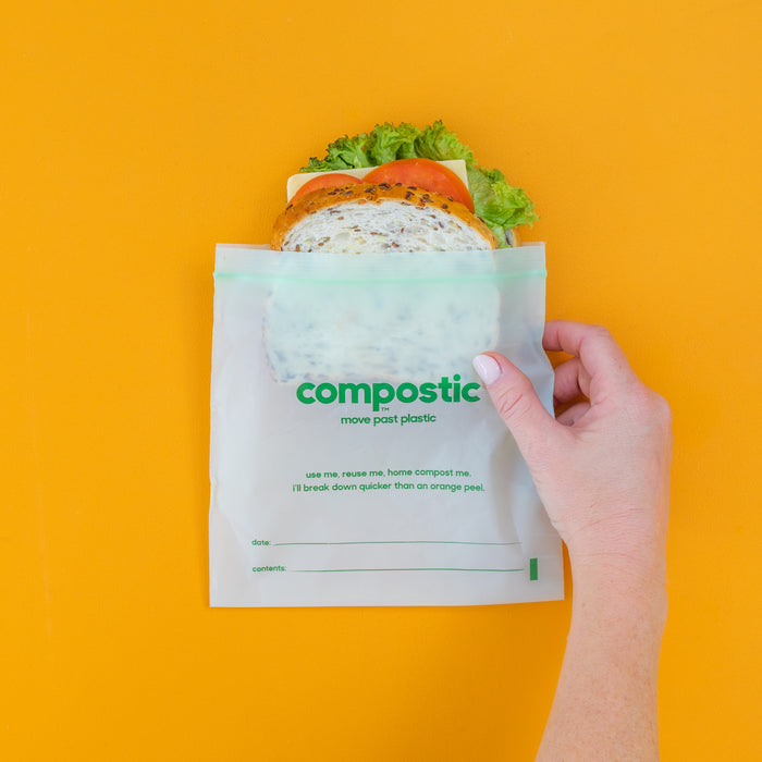 A hand holds a sandwich in a Compostic Sandwich Bag, made from home-compostable, plant-based materials. Against an orange backdrop, the bag promotes composting and sustainability.