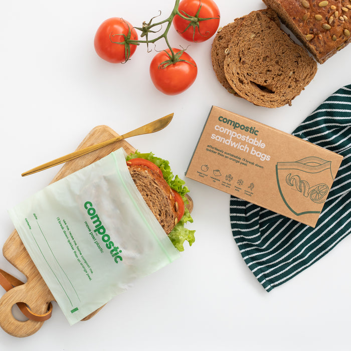 A sandwich with lettuce and tomato is in a Compostic Sandwich Bag on a wooden board. Nearby, sliced bread, fresh tomatoes, a knife, and a box labeled Compostic Sandwich Bags are on a striped cloth.