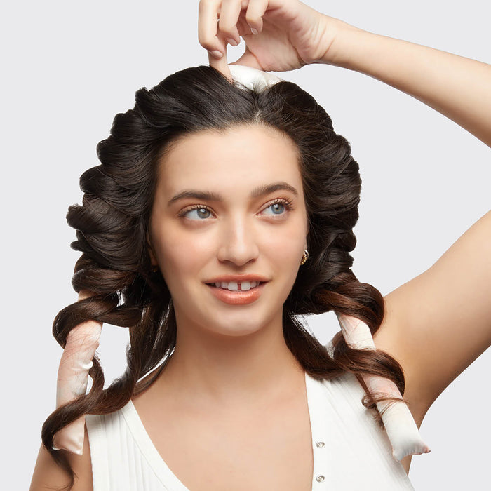 With their long, dark hair styled in large curls, a person smiles and touches their head, enjoying frizz-free hair thanks to the Kitsch Satin Heatless Curling Set. Theyre wearing a white sleeveless top against a plain background.
