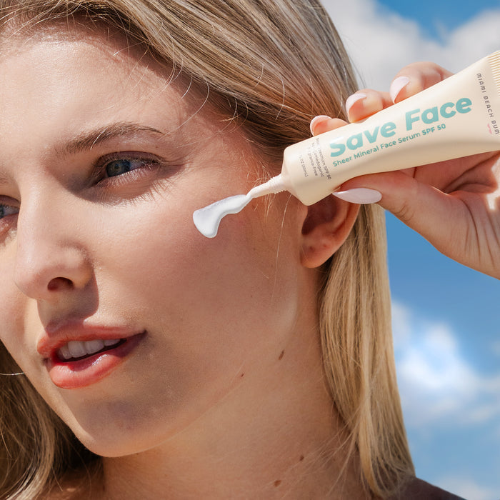 A fair-skinned person with light hair holds a tube labeled Save Face Serum - SPF50 near their cheek, squeezing out sheer mineral cream. This Miami Beach Bum non-comedogenic serum is Hawaii Reef-Friendly. The clear background highlights the products elegance.