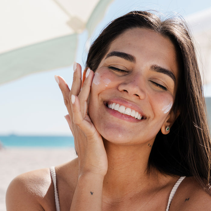 A woman with long dark hair and a hand tattoo smiles as she applies Miami Beach Bums Save Face Serum - SPF50. Outdoors at the beach under a sunshade, the reef-friendly formula keeps both her skin and the ocean protected against the backdrop of blue sea and sky.